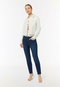 Biała dżinsowa kurtka z guzikami, stylizowana na beżowej koszulce. Ciemnoniebieskie slim-fit jeansy i białe sandały na słupku dopełniają całość.