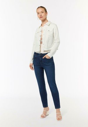 Veste en denim blanche avec des boutons, portée sur un haut beige. Un jeans slim-fit bleu foncé et des sandales à talons blocs blanches à lanières complètent le look.