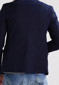 Navy blue tailored blazer with a smooth texture, featuring a single back vent and structured shoulders, paired with denim jeans.