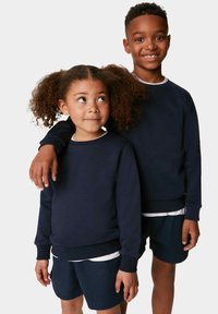 Dos niños están de pie uno al lado del otro, vistiendo sudaderas oscuras azules y pantalones cortos a juego. La tela parece suave, con puños y dobladillos acanalados.