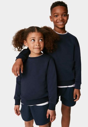 Dos niños están de pie uno al lado del otro, vistiendo sudaderas oscuras azules y pantalones cortos a juego. La tela parece suave, con puños y dobladillos acanalados.