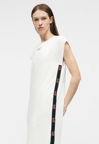 Γυναίκα με μακριά καστανά μαλλιά που φοράει ένα λευκό αμάνικο φόρεμα με μαύρη κάθετη ρίγα και πορτοκαλί λογότυπα EA7 στο πλάι.