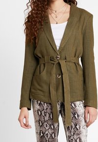 Olivově zelený sako s opaskem vyrobené z texturované tkaniny, se zapínáním na jeden knoflík a dvěma předními kapsami. V páru s kalhotami s hadím potiskem.