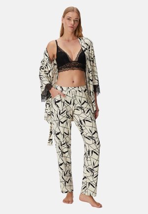 Vrouw poseert in een zwarte kanten bralette en een wit-zwart patroon pyjama set met kamerjas, staand op blote voeten tegen een effen achtergrond.