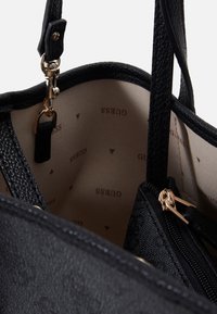 Close-up van de binnenkant van een zwarte leren handtas met een beige stofvoering bedrukt met "GUESS" en gouden rits- en sluitdetails.