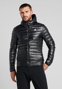 Schwarze Pufferjacke mit Kapuze, mit einem Reißverschluss vorne, horizontalen Steppnähten und einem kleinen weißen Logo auf der linken Brust. Glattes, glänzendes Material.