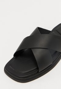 Svart slip-on sandal med kryssreimer laget av glatt skinn, med en flat rektangulær såle og subtil merking på siden.