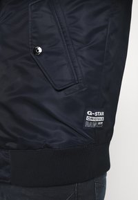Marinblå bomberjacka tillverkad av ett slätt tyg. Har ett märke med "G-Star RAW"-logotyp och en knappad ficka med lock. Ribbad mudd på ärmarna.