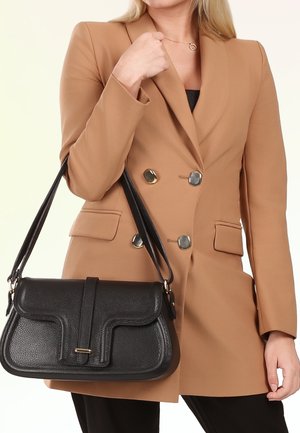 Vrouw in een camelkleurige blazer houdt een zwarte leren schoudertas met een getextureerd klepje en een gouden gesp vast.