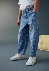 Pantaloni din denim cu croială lejeră, culoare albastru deschis și buline mari albe. Asortați cu adidași albi, evidențiind un look casual.