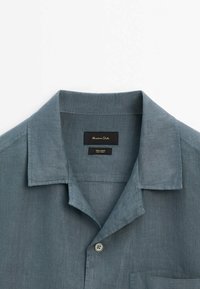 Chemise bleue en lin avec col, boutonnée, un seul bouton visible et une poche poitrine, étiquette intérieure du col indiquant "100% lin".