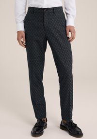 Muž v tmavých vzorovaných kalhotách, bílém tričku, béžových ponožkách a černých kožených polobotkách na neutralním pozadí.