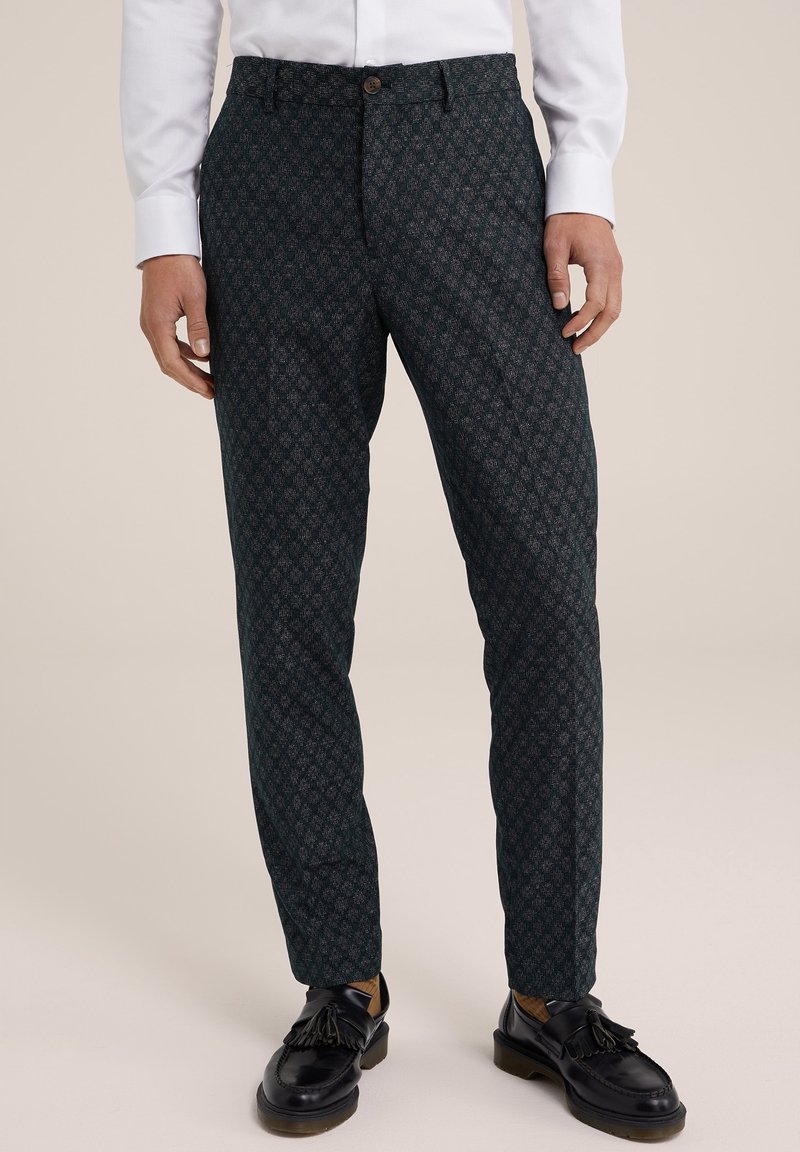 Muž v tmavých vzorovaných kalhotách, bílém tričku, béžových ponožkách a černých kožených polobotkách na neutralním pozadí.