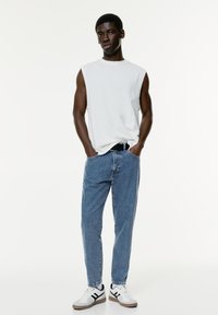 Biała bezrękawnikowa bluzka, niebieskie dżinsy i białe sneakersy z czarnymi akcentami. Slim-fit sylwetka o casualowym wyglądzie.