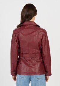 Kvinde iført en tætsiddende bordeaux læderjakke med bælte, lynlåslukkede ærmer og blå jeans, vist fra ryggen mod en ensfarvet baggrund.