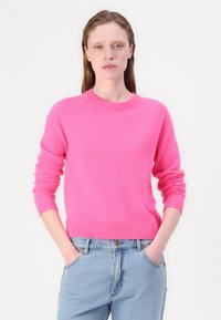 Lys rosa strikkegenser med rund hals, ribbestrikkede ermer og kant, brukt over lyseblå jeans. Myk tekstur, avslappet passform.