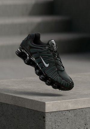 Čierna a tmavosivá bežecká obuv Nike s bielym logom swoosh zavesená nad betónovým okrajom a schodmi v pozadí.