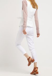 Biała bluzka z baskinką nałożona na jasnoróżową bluzkę z długim rękawem, zestawiona z białymi zwężanymi spodniami i brązowymi sandałami na obcasie.