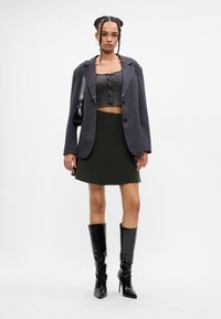 Žena s vrkôstím nosí šedý oversized blazer, rebrovaný crop top, tmavú sukňu, čiernu kabelku a čierne kožené čižmy po kolená.