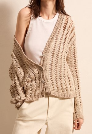 Femme portant un top blanc sans manches sous un cardigan beige en maille ajourée avec des boutons et un style épaules dénudées, associé à un pantalon crème.