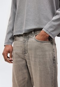 Άτομο που φοράει τζιν παντελόνι ανοιχτού γκρι και μακρυμάνικο γκρι πουκάμισο, με το δεξί χέρι στην μπροστινή τσέπη του τζιν.