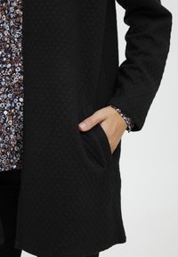 Giacca nera in tessuto con vestibilità ampia, dotata di una tasca frontale e una camicia a fiori con motivo che spunta dalla manica.