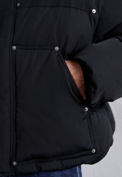 Hand in der Tasche einer schwarzen gepolsterten Jacke mit Druckknöpfen an den Taschenrändern, vor einem neutralen Hintergrund gezeigt.