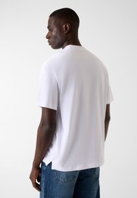 Biały bawełniany t-shirt z krótkimi rękawami, luźnym krojem i bocznymi rozcięciami. Noszony z niebieskimi jeansami, widoczny od tyłu. Gładka faktura.