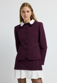 Blazer violet avec un col Peter Pan blanc, des boutons sur le devant et deux poches. Accompagné d'une jupe blanche à volants. Texture lisse.