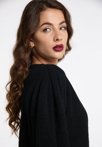 Maglione nero lavorato a maglia con un motivo testurizzato e sottili maniche a sbuffo, indossato con capelli lunghi e ondulati e rossetto bordeaux audace.