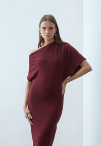 Burgundijas krāsas kleita bez pleca ar brīvu piegriezumu, izgatavota no gludas auduma, kas eleganti pieguļ ķermenim. Garas piedurknes, minimāla dizaina.