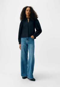 Donna con capelli ricci, che indossa una blusa nera a maniche lunghe e jeans blu a gamba larga, in piedi con una mano in tasca.