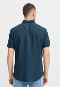 Chemise à manches courtes en tissu bleu foncé, dotée d'un col standard et d'un ourlet arrondi. Fini simple et texturé, sans motifs ni accents visibles.