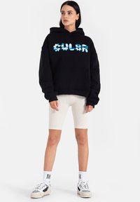 Czarna bluza z kapturem z kolorowym nadrukiem tekstowym, z kieszenią na przodzie i długimi rękawami. W zestawie z jasnymi, prążkowanymi szortami rowerowymi i trampkami.