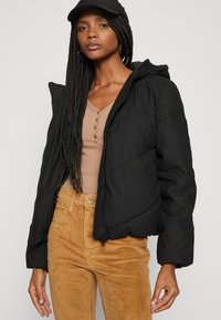 Veste noire rembourrée avec capuche, dotée d'une fermeture éclair à l'avant et de poignets côtelés, portée sur un haut beige et un pantalon en velours côtelé couleur tan.