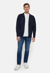 Marineblaues Zip-Jackett mit hohem Kragen, kombiniert mit einem weißen T-Shirt und blauen Jeans. Verfügt über zwei Seitentaschen und gerippte Bündchen.