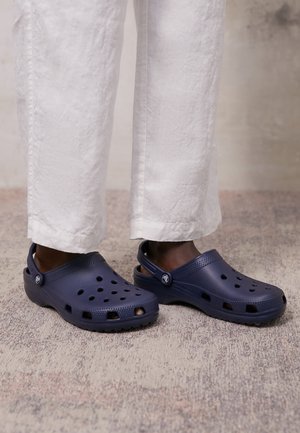 Personne portant des sabots bleu foncé à enfiler avec des trous de ventilation et des brides réglables au talon, associés à un pantalon court blanc sur un sol texturé.