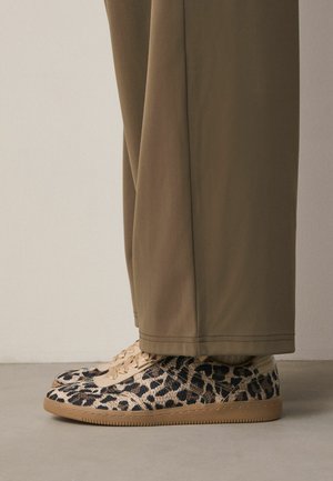 Niedrige Sneaker mit Leopardenmuster, beigen Sohlen und Schnürsenkeln, getragen zu lockeren olivgrünen Hosen, stehend auf einem grauen Boden.
