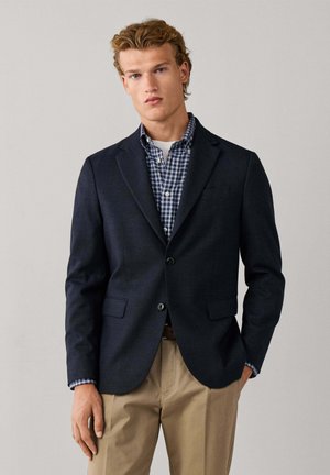 Young man with curly blond hair wears navy blazer, blue and white checkered shirt, and beige trousers, standing against plain light background.