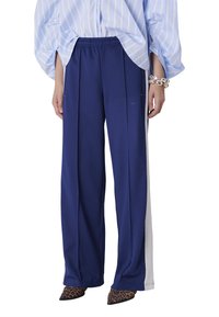 Pantalon bleu marine avec ceinture élastique, doté de bandes latérales blanches et de détails plissés. Associé à une chemise rayée bleu clair et des chaussures à imprimé léopard.