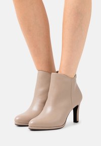 Stivali alla caviglia in pelle beige con una trama liscia, punte appuntite e un bordo superiore curvo, dotati di un tacco alto e sottile di colore nero.