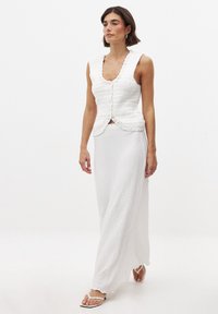 Valge kudum vest ilma varrukateta, millel on laineline kaelus ja nööp sulgemine, koos pika valge tekstuuriga seelikuga, kantud valgete sandaalidega.