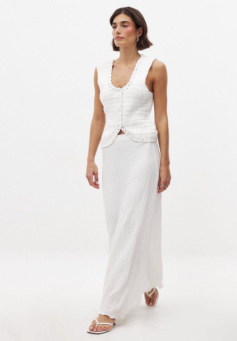 Valge kudum vest ilma varrukateta, millel on laineline kaelus ja nööp sulgemine, koos pika valge tekstuuriga seelikuga, kantud valgete sandaalidega.