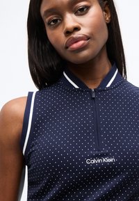 Νεαρή γυναίκα που φοράει αμάνικη μπλε μπλούζα με λευκές πουά και λογότυπο Calvin Klein στο μπροστινό μέρος, με φερμουάρ στον γιακά.