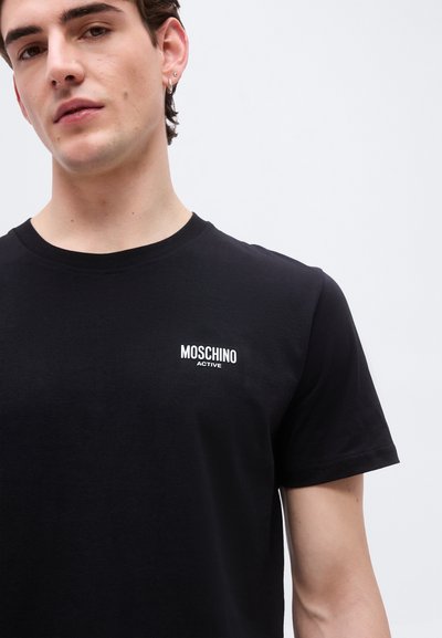 Νεαρός άνδρας φοράει μαύρο μπλουζάκι Moschino Active με μικρό λευκό λογότυπο στο στήθος, στέκεται μπροστά σε απλό ανοιχτό φόντο.