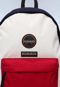 Sac à dos rouge et blanc au design arrondi, avec un logo noir circulaire et une étiquette rectangulaire portant le nom de la marque. Texture de tissu durable.