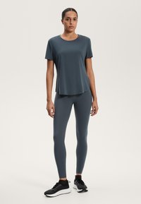 Sötétszürke, nedvességet elvezető, rövid ujjú póló, amelyet ugyanolyan színű, szoros leggings-szel és fekete sportcipővel párosítottak, semleges háttér előtt.