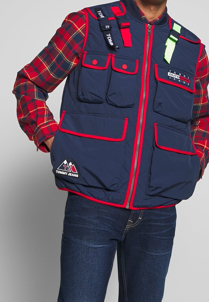 Homme portant un gilet bleu marine avec bordure rouge et multiples poches par-dessus une chemise à carreaux rouge et un jean bleu, debout les mains dans les poches.