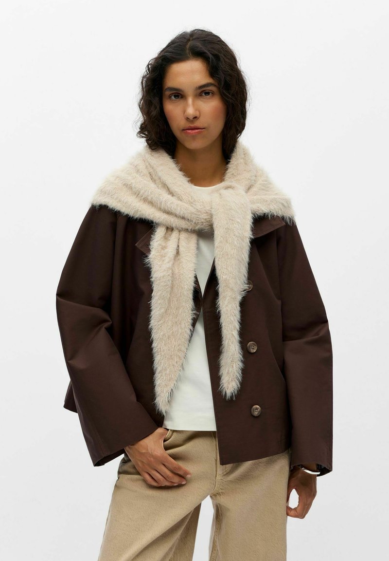 Jeune femme aux cheveux bouclés porte une veste marron foncé, un pantalon beige, un haut blanc et une écharpe crème duveteuse drapée autour de ses épaules.