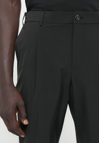 Svarta byxor i lätt tyg med en slät textur. Funktioner inkluderar en knappstängning, sidofickor och plisserade detaljer.